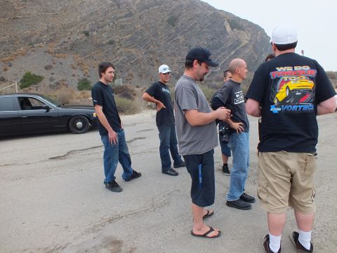 2003 Ford Crown Victoria - group of men in t-shirts and jeans stand talking outdoors beside a black sedan with a rocky hillside behind them.
