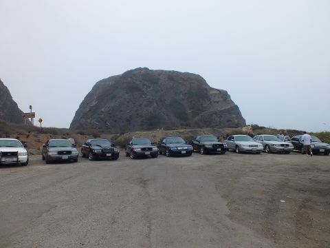 2003 Ford Crown Victoria - Front-facing full-size sedans parked across a wide gravel lot beneath a large dome-shaped rock in foggy light.