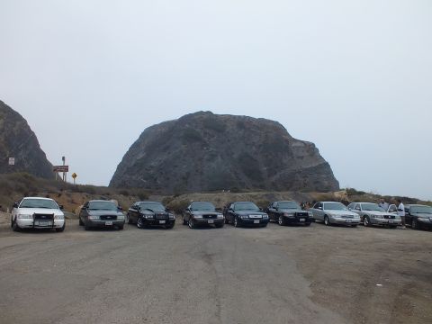 2003 Ford Crown Victoria - Front-facing full-size sedans line up in a gravel lot below a large rock formation under overcast gray sky.