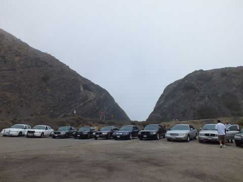 2003 Ford Crown Victoria - row of white and black full-size sedans parked in a gravel lot in the gap between two rocky hills, with a person at right.