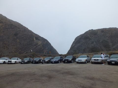 2003 Ford Crown Victoria - lineup of dark and silver full-size Ford sedans parked across a gravel coastal lot under heavy fog between two hills.