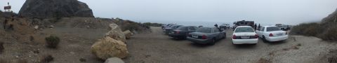 2003 Ford Crown Victoria - wide panoramic shot of sedans parked on a rocky gravel pull-out beside the ocean, framed by boulders and a foggy headland.
