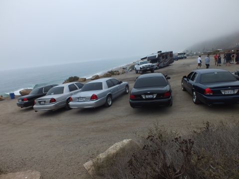 2003 Ford Crown Victoria - Black and silver full-size Ford sedans parked at an angle on a coastal bluff while people gather near an RV by the foggy ocean.