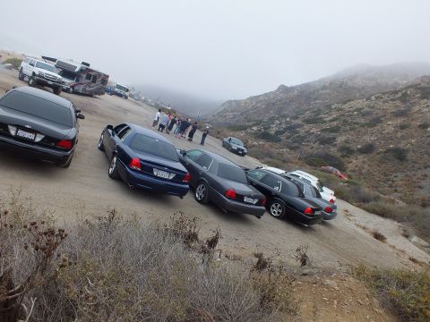 2003 Ford Crown Victoria - cluster of dark blue and black Ford sedans parked on a dirt slope as people gather near an RV beneath a foggy ridgeline.
