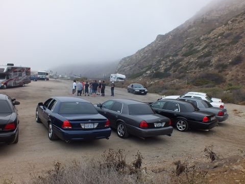 2003 Ford Crown Victoria - Dark full-size Ford sedans parked in a loose semicircle on a gravel coastal lot while people stand together near an RV under fog.