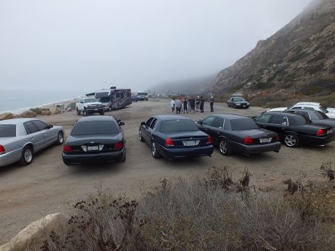 2003 Ford Crown Victoria - lineup of silver and dark Ford sedans on a foggy gravel coastal lot, with a small crowd and an RV near the shoreline.