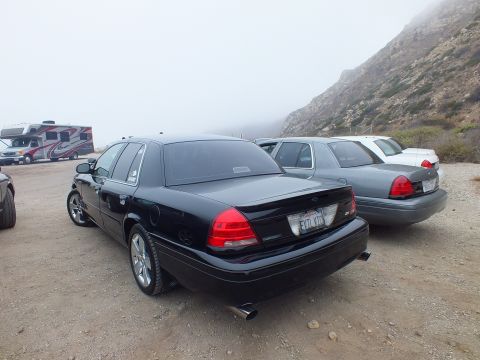 2003 Ford Crown Victoria - Rear three-quarter view of a black full-size Ford sedan with a vanity plate reading EVIL VIC, parked beside a silver sedan on gravel.
