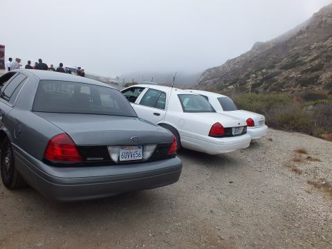 2003 Ford Crown Victoria - Rear of a gray full-size Ford sedan with plate 6UVV656 parked beside two white sedans on a gravel lot below a foggy hill.