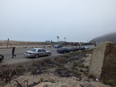 2003 Ford Crown Victoria - silver sedan and several dark cars travel along a coastal highway beside dunes and a boulder under thick gray fog.