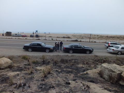 2003 Ford Crown Victoria - Two black sedans pulled onto a coastal highway shoulder as people stand between them, with a beach and parking area beyond.