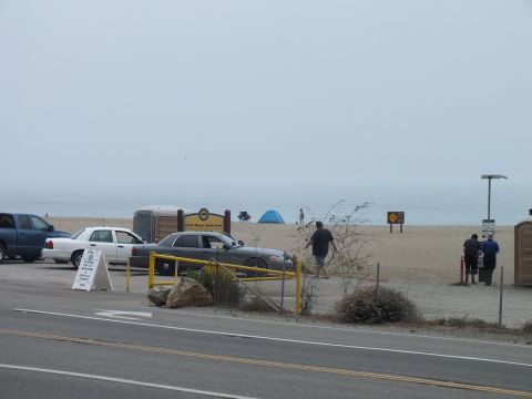 2003 Ford Crown Victoria - state beach park entrance with a yellow gate, portable restroom and sign as sedans park near the sand under gray fog.