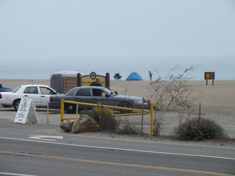 2003 Ford Crown Victoria - beach park entry gate with white Ford sedans and a portable toilet near the road, a tent pitched on the sand beyond.