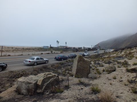 2003 Ford Crown Victoria - silver sedan and dark cars travel a coastal highway seen across a foreground of boulders and dry brush under fog.