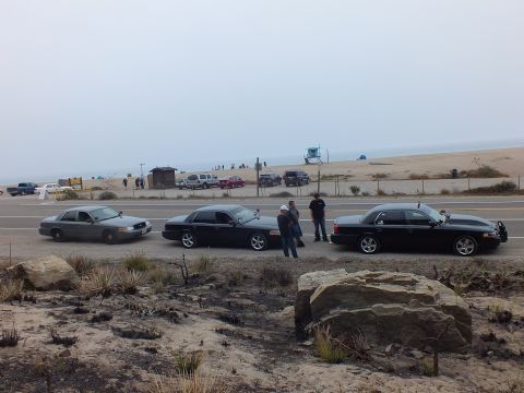 2003 Ford Crown Victoria - Three dark sedans are parked roadside near a foggy beach as a few people stand between them.