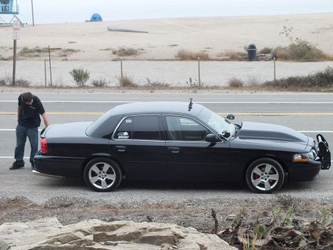 2003 Ford Crown Victoria - lowered black full-size sedan is shown in side profile beside a beach while a man stands near its trunk.