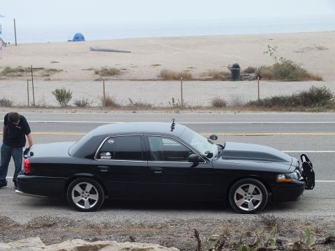 2003 Ford Crown Victoria - lowered black full-size sedan sits in profile by a fenced beach with a person beside the rear.