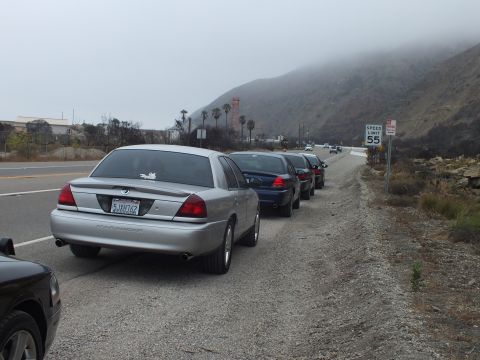 2003 Ford Crown Victoria - silver Mercury sedan leads a line of cars pulled onto a highway shoulder below a fog-capped hill.