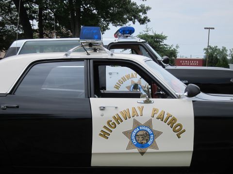 1991 Ford LTD Crown Victoria - Close-up of a black-and-white police car's front door bearing the gold California Highway Patrol star emblem, with blue roof lights above.