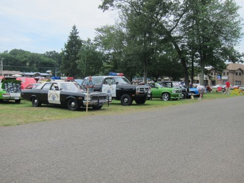 1991 Ford LTD Crown Victoria - row of vehicles including black-and-white patrol cars and a green muscle car lined up along pavement at an outdoor car show.