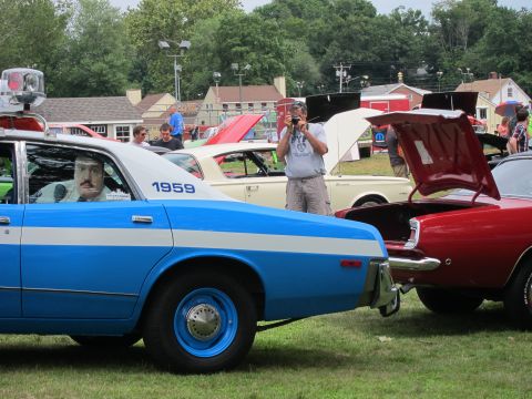 1991 Ford LTD Crown Victoria - blue-and-white police sedan numbered 1959 with a uniformed mannequin inside, parked beside a red car as a photographer shoots nearby.