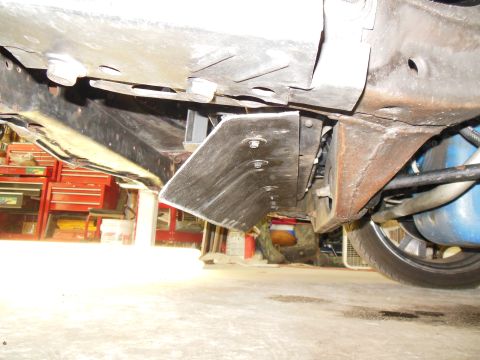 1979 Ford Ranchero - Low ground-level view under the front of a car on a garage floor, showing a bare steel frame rail, floor pan and a flat bolt-on bracket pla…