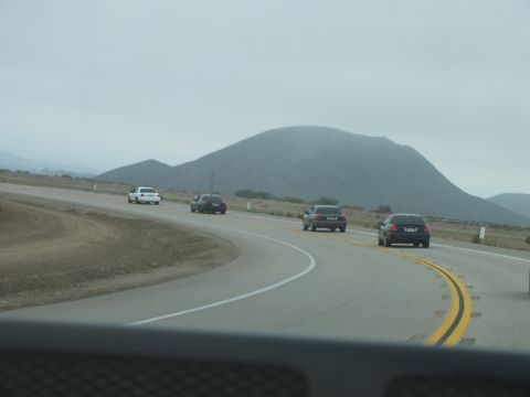 2003 Ford Crown Victoria - Seen through a windshield, dark sedans round a highway curve toward a large barren rounded hill.
