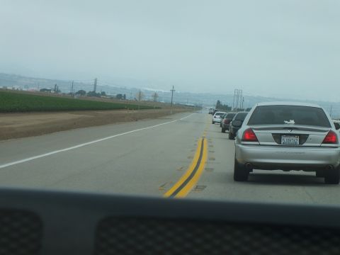 2003 Ford Crown Victoria - rear of a silver Mercury Marauder follows a line of cars on a highway beside green farm fields.