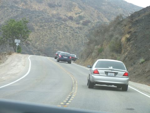 2003 Ford Crown Victoria - silver sedan and dark cars climb a winding two-lane road through dry brushy mountain terrain.