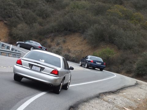 2003 Ford Crown Victoria - silver sedan and two dark sedans round a tight bend on a road through green brushy hills.