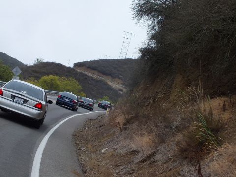 2003 Ford Crown Victoria - silver sedan trails dark sedans along a guardrail-lined road through dry hills near a power tower.