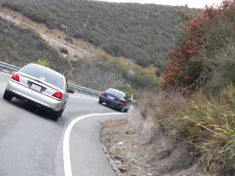 2003 Ford Crown Victoria - silver sedan and a dark sedan descend a curving guardrail-lined road through brushy hills.