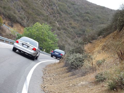 2003 Ford Crown Victoria - silver sedan and a dark sedan follow a curving guardrail-lined road across a steep brushy slope.