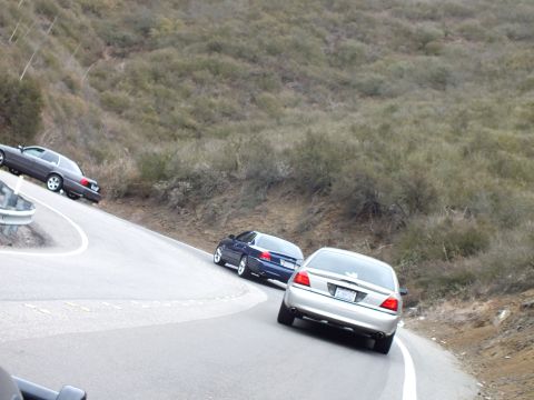 2003 Ford Crown Victoria - silver sedan and dark cars navigate a tight switchback on a road through dry brushy hills.