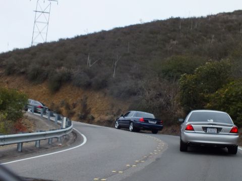 2003 Ford Crown Victoria - silver sedan and a dark sedan travel a winding road below a brushy hillside near a power tower.