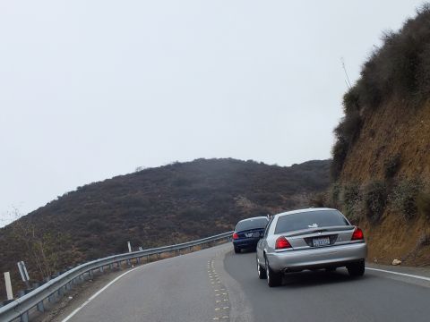 2003 Ford Crown Victoria - silver full-size sedan trails a dark sedan along a curving foggy two-lane mountain highway lined by a guardrail and brushy hillside.