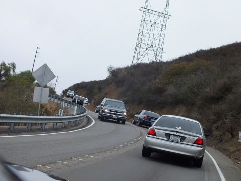 2003 Ford Crown Victoria - silver full-size sedan and a line of vehicles wind down a curved hillside road under a large steel transmission tower on a gray day.