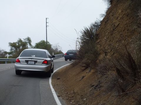 2003 Ford Crown Victoria - silver full-size sedan and a dark sedan sit along a road edge beneath a steep eroded dirt embankment under an overcast sky.