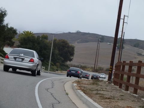 2003 Ford Crown Victoria - silver full-size sedan leads a line of dark sedans pulled over on a rural roadside with golden hills and a wooden fence behind.
