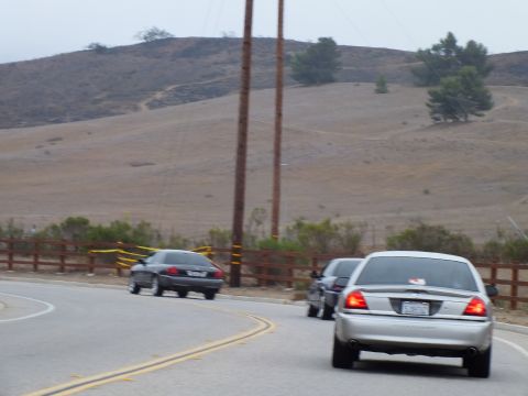 2003 Ford Crown Victoria - silver full-size sedan and a dark sedan drive a curving two-lane road below bare golden hills and tall utility poles.