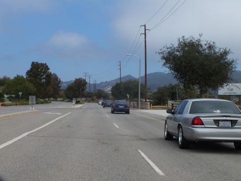 2003 Ford Crown Victoria - silver full-size sedan travels a wide multi-lane road toward distant blue mountains, with other cars and a roadside sign ahead.