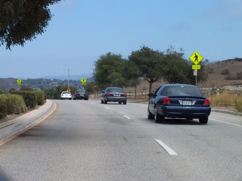 2003 Ford Crown Victoria - dark blue full-size sedan and other cars travel a curving road past a yellow pedestrian-crossing sign and a low wooden fence.