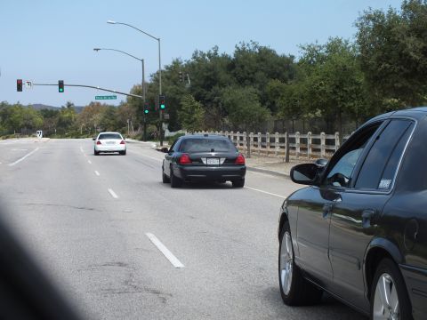 2003 Ford Crown Victoria - Dark full-size sedans wait and proceed through a green traffic light on a tree-lined road, with a wooden fence along the shoulder.