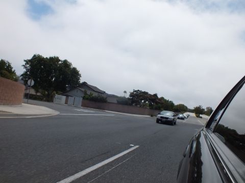 2003 Ford Crown Victoria - dark sedan crosses a wide intersection near suburban homes and a brick wall, photographed from another car under heavy clouds.