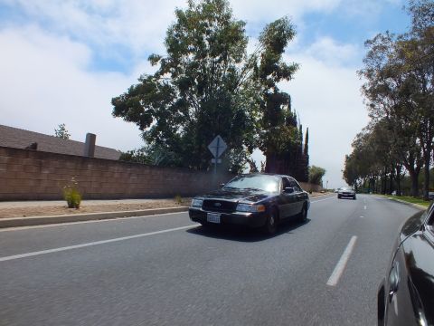 2003 Ford Crown Victoria - black Ford full-size sedan approaches on a multi-lane road past a brick wall, tall trees, and a diamond road sign.