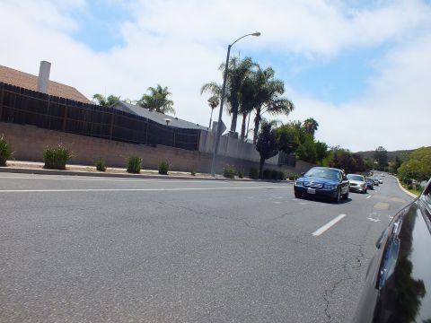 2003 Ford Crown Victoria - dark blue full-size sedan leads a line of cars along a wide road beside a long building, fence, and clustered palm trees.