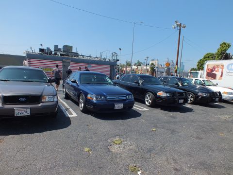 2003 Ford Crown Victoria - Several Ford full-size sedans in silver, blue, and black are parked side by side in an urban lot near a building and a box truck.