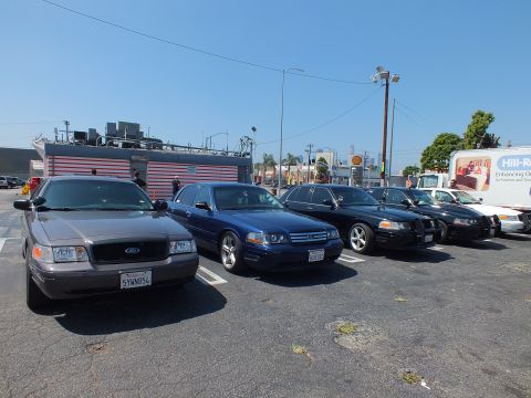 2003 Ford Crown Victoria - lineup of Ford full-size sedans in silver, dark blue, and black sits angled in a paved urban lot with a building and box truck behind.