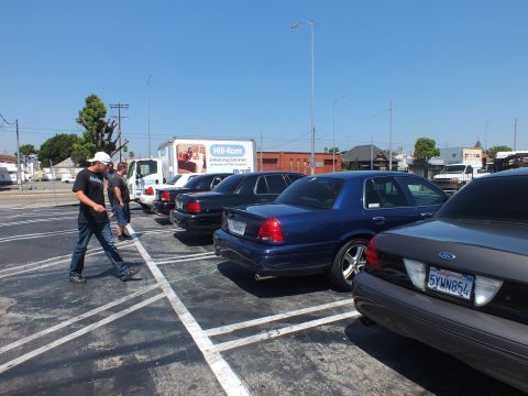2003 Ford Crown Victoria - People walk past a row of parked dark Ford full-size sedans in a paved lot beside a Hill-Rom box truck under a clear blue sky.