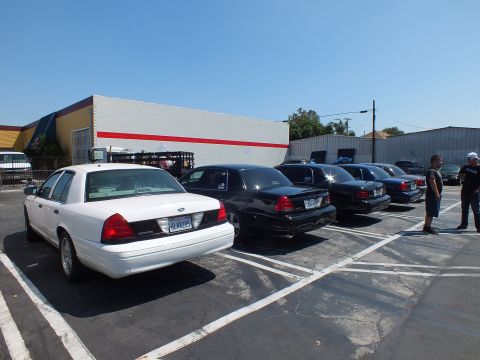 2003 Ford Crown Victoria - white Ford full-size sedan and a row of black sedans sit parked in a lot beside a white building with a red stripe; two men stand at right.