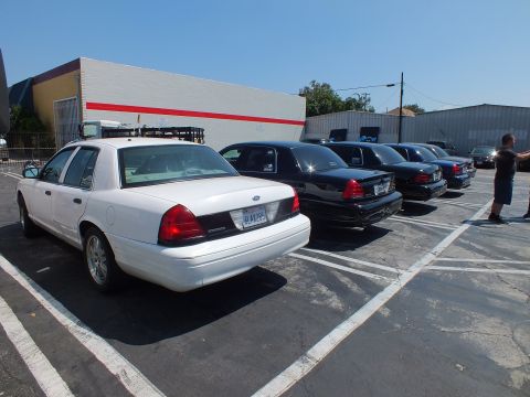 2003 Ford Crown Victoria - white Ford full-size sedan and a line of black sedans are parked in marked stalls beside a white, red-striped building, a man standing near…